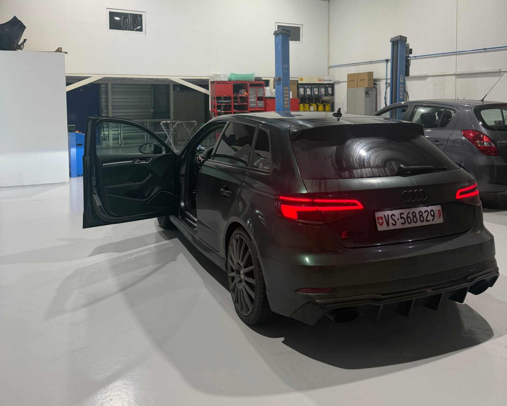 Une Audi à hayon gris foncé, portière ouverte, garée dans un atelier automobile baigné de lumière.