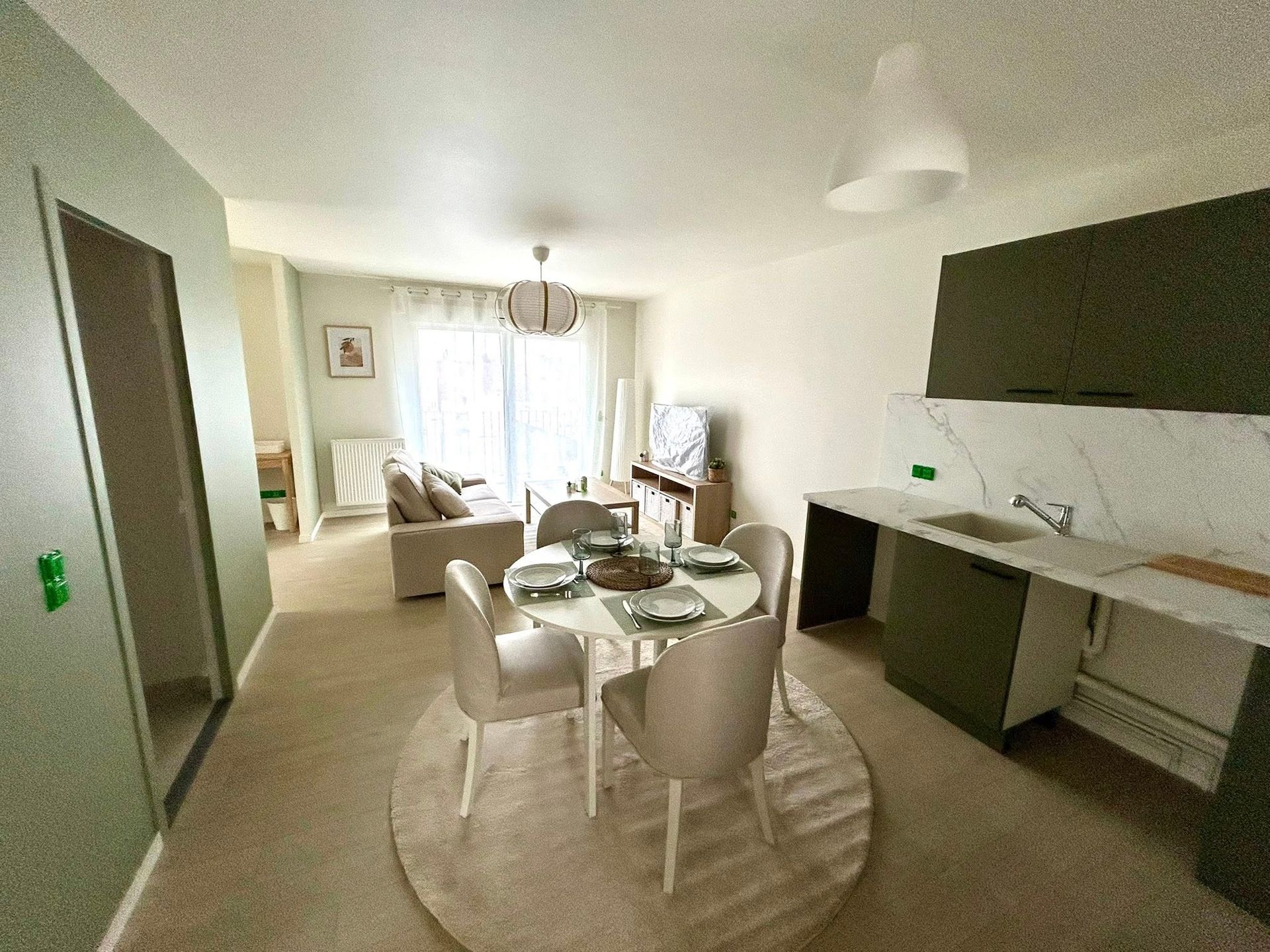 Un petit appartement avec un coin repas, une cuisine et un salon. Murs vert, mobilier aux tons neutres.