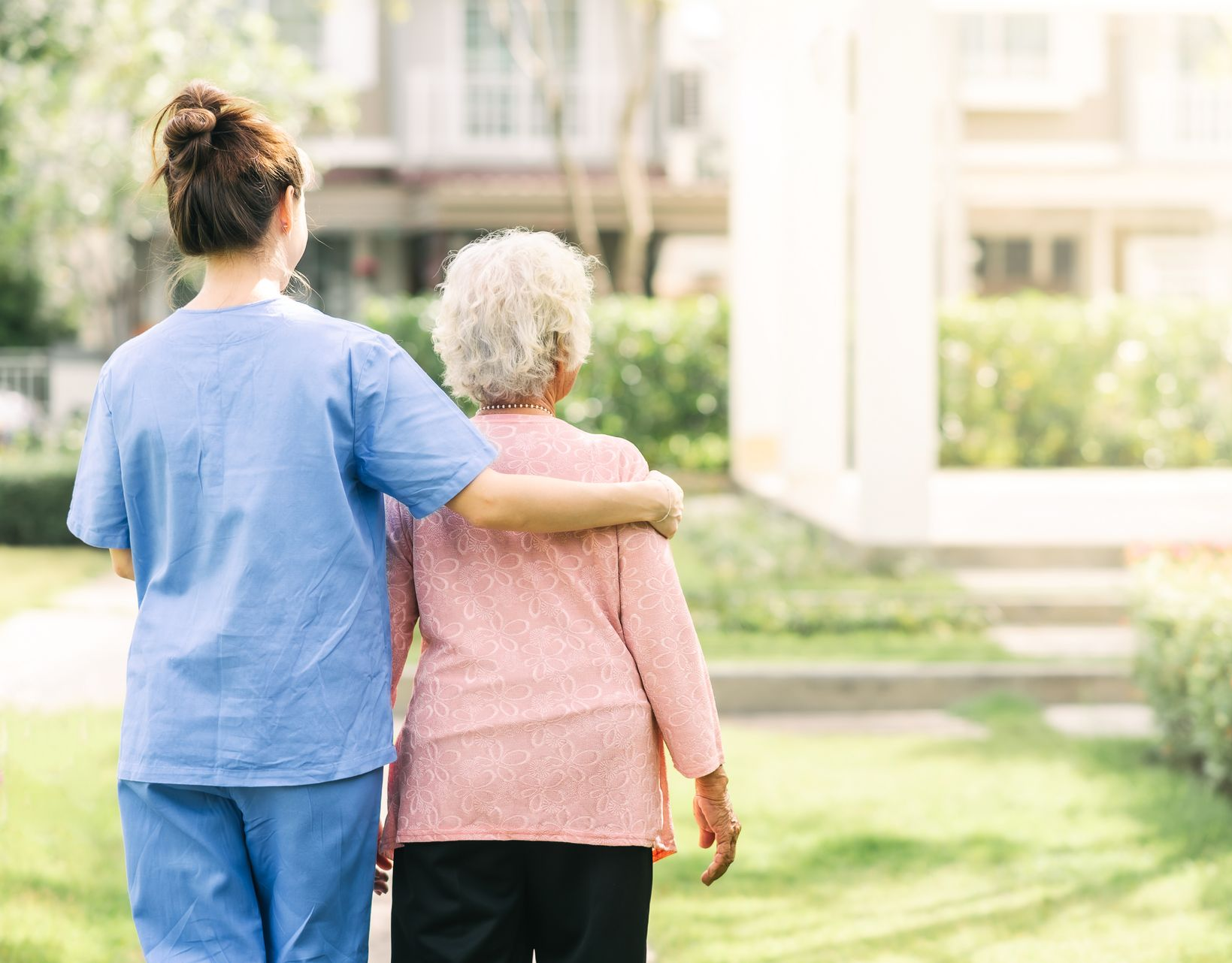 Une aide-soignante en blouse bleue accompagne une femme âgée qui marche à l'extérieur.