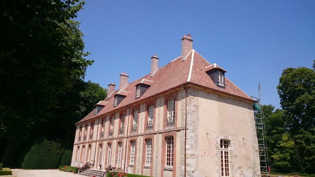 Manoir historique en pierre rose avec toit et cheminées rouges, vu de côté dans un jardin ensoleillé