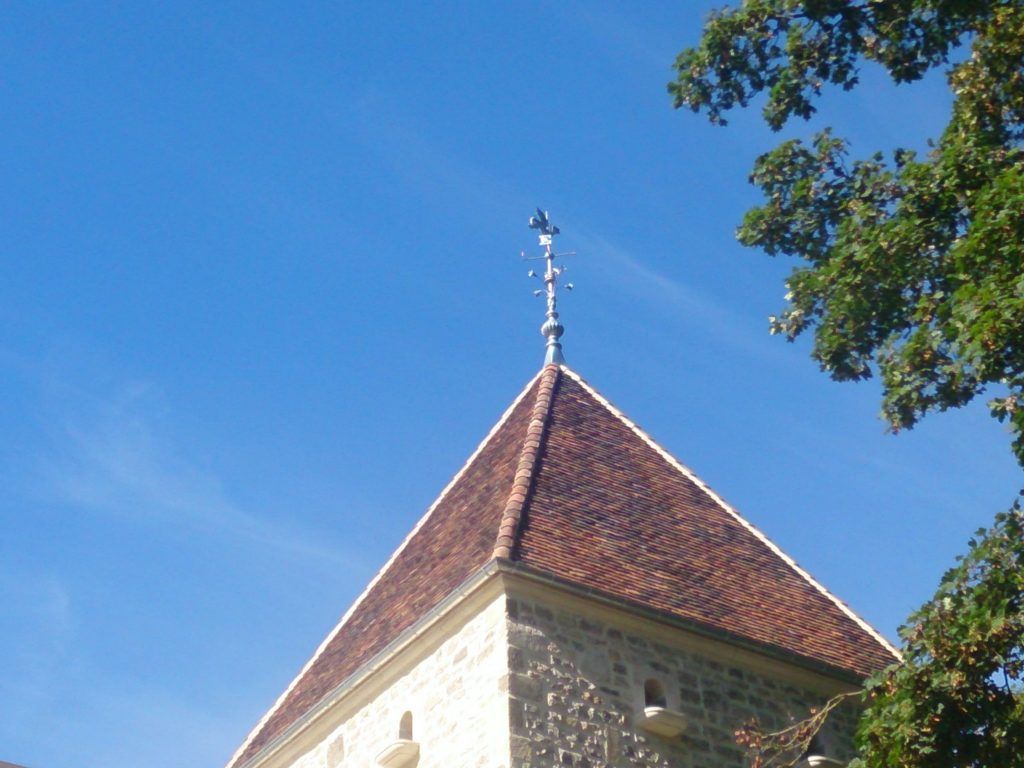 Clocher en pierre avec un toit de tuiles rouges et une girouette se détachant sur un ciel bleu, encadré par des branches d'arbres