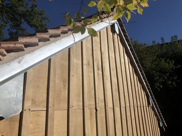Mur d'abri en bois avec planches verticales et moulures blanches sur le toit, se détachant sur un ciel bleu avec des branches au-dessus.