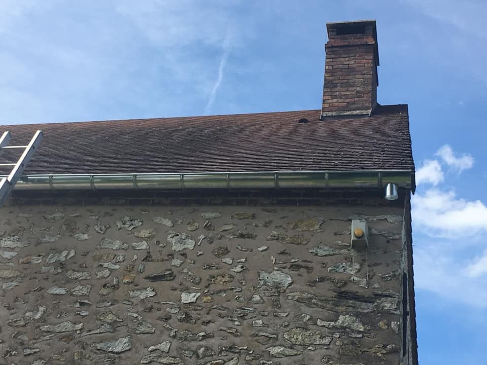 Mur en pierre, toit sombre et cheminée en briques se détachant sur un ciel bleu.