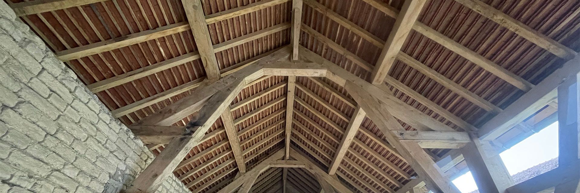 Vue intérieure d'un toit de chaume avec poutres en bois et mur en pierre, éclairé par des puits de lumière.