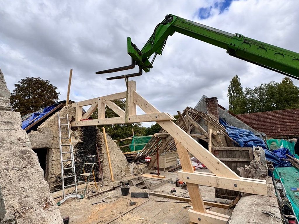 Une excavatrice soulève une charpente en bois pour la déposer sur un bâtiment en pierre en construction, avec des bâches bleues et des gravats à proximité.