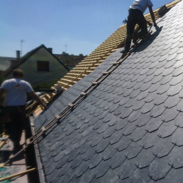 Des ouvriers posent des bardeaux sombres sur le toit pentu d'une maison, sous un ciel bleu.