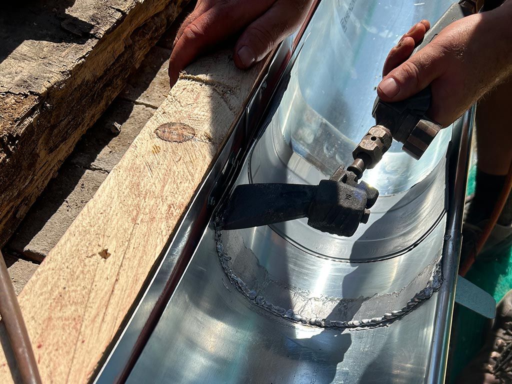 Des mains appliquent du mastic sur un tuyau de cheminée métallique brillant, sur un toit.