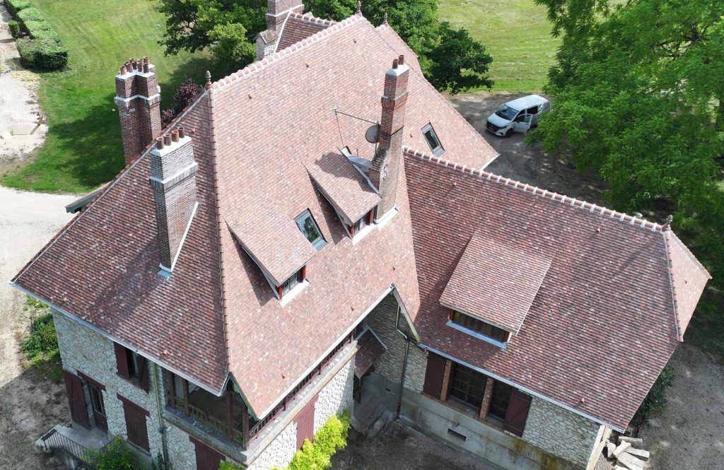 Vue aérienne d'une maison en pierre avec un toit de tuiles rouges et des lucarnes, entourée d'arbres verts.