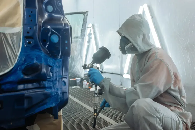 Una persona con traje de protección rocía pintura azul sobre el chasis de un automóvil en una cabina de pintura.