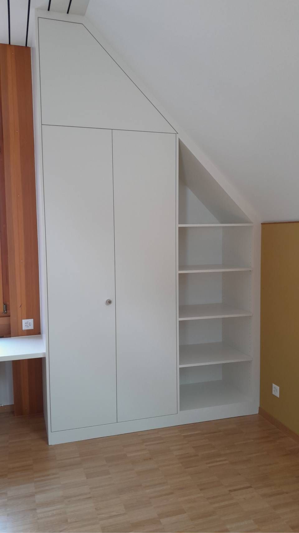 Weisser, eingebauter Dachbodenschrank mit Regalen und geschlossenen Türen neben einer Dachschräge in einem Zimmer mit Holzboden