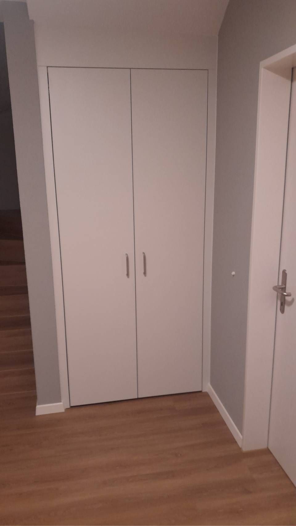 Weisse Doppelschranktüren in einem kleinen Flur mit hellem Holzboden und einer Tür auf der rechten Seite.