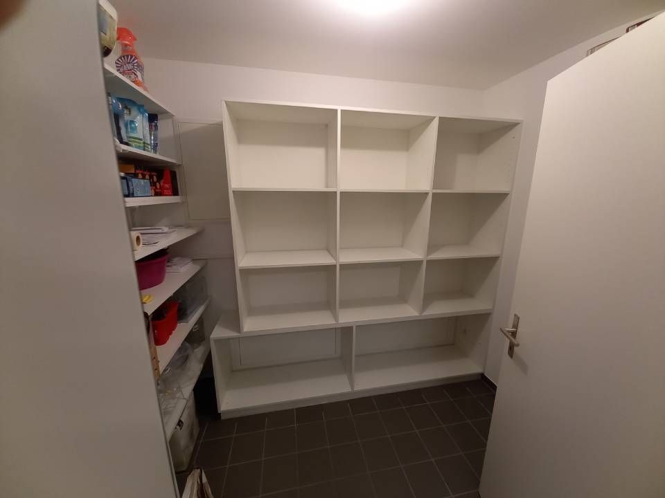 Leere weisse Einbauregale in einem kleinen Schrank mit dunklem Fliesenboden und Reinigungsmitteln auf der linken Seite.
