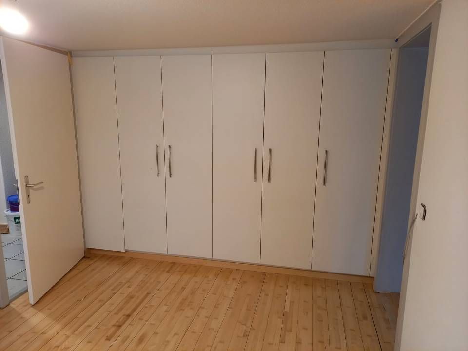 Grosszügiges, leeres Zimmer mit weissen Kleiderschränken und hellem Holzboden, gesehen von einer Türöffnung aus.