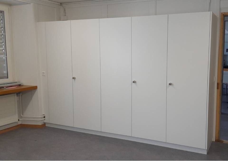 Weisse Aufbewahrungsschränke säumen eine Wand in einem schlichten Zimmer mit grauem Teppichboden und einem kleinen Fenster.