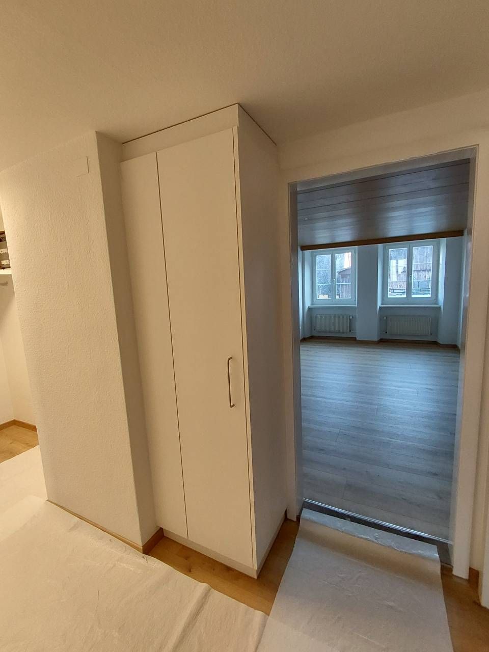 Flur mit einem weissen Schrank neben einer offenen Tür zu einem hellen Zimmer mit Fenstern und Holzboden