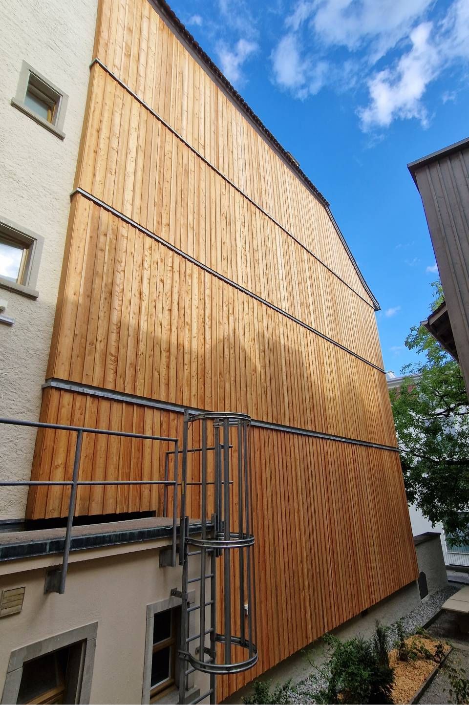 Eine schmale Gasse zwischen zwei Gebäuden, mit einer hohen, holzverkleideten Fassade und blauem Himmel darüber.