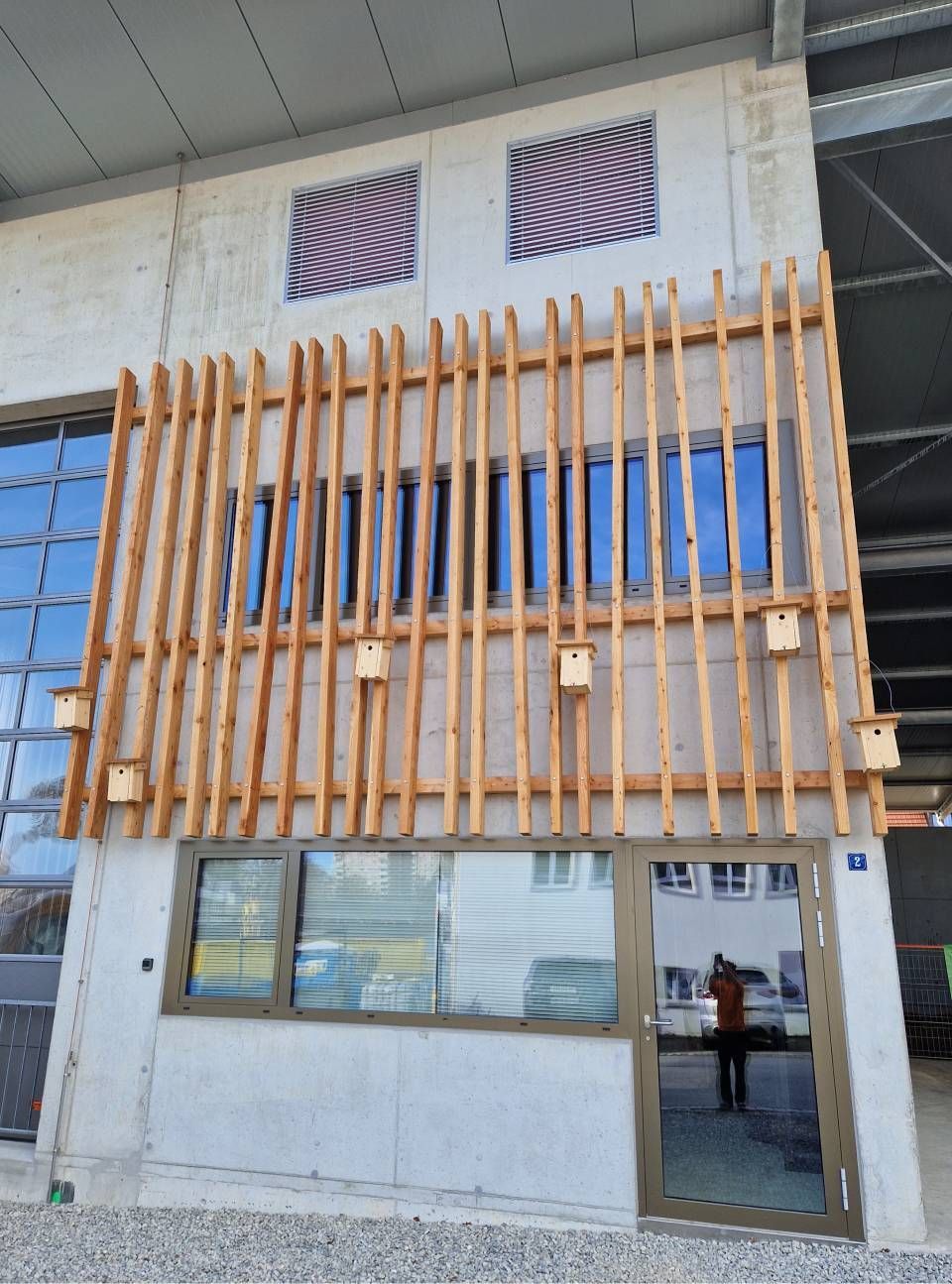 Moderne Gebäudefassade mit vertikalen Holzlamellen, grossen Fenstern und einem gläsernen Eingang.