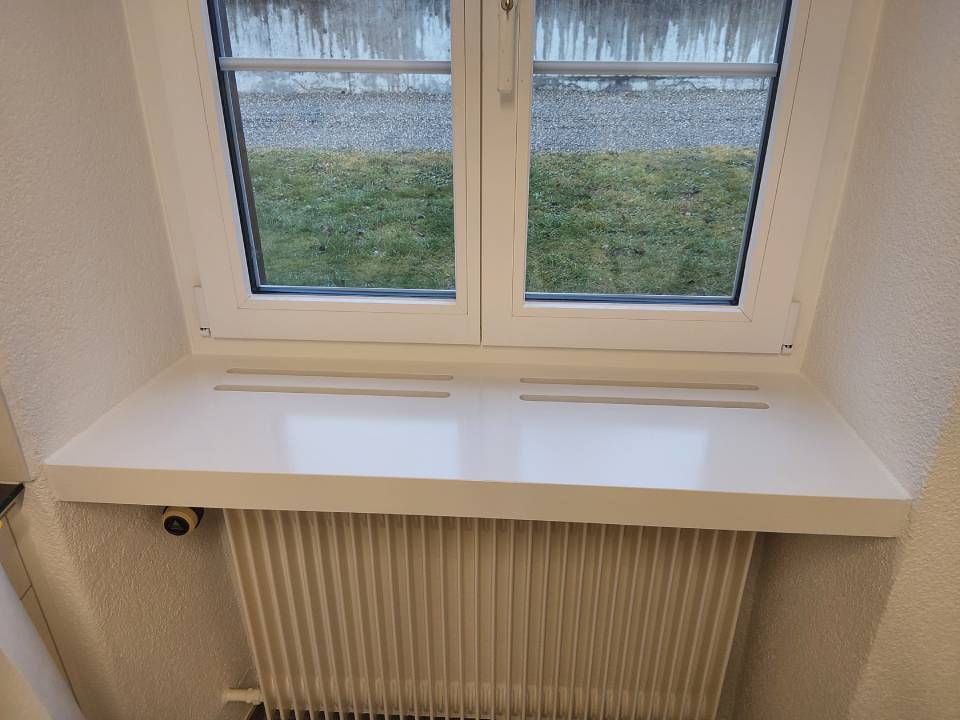 Weisses Fensterbrett über einem beigen Heizkörper unter einem kleinen Fenster, draussen ist Gras zu sehen.