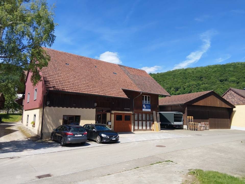 Ländliches Haus mit rotem Ziegeldach, geparkte Autos und grüner Hang unter blauem Himmel