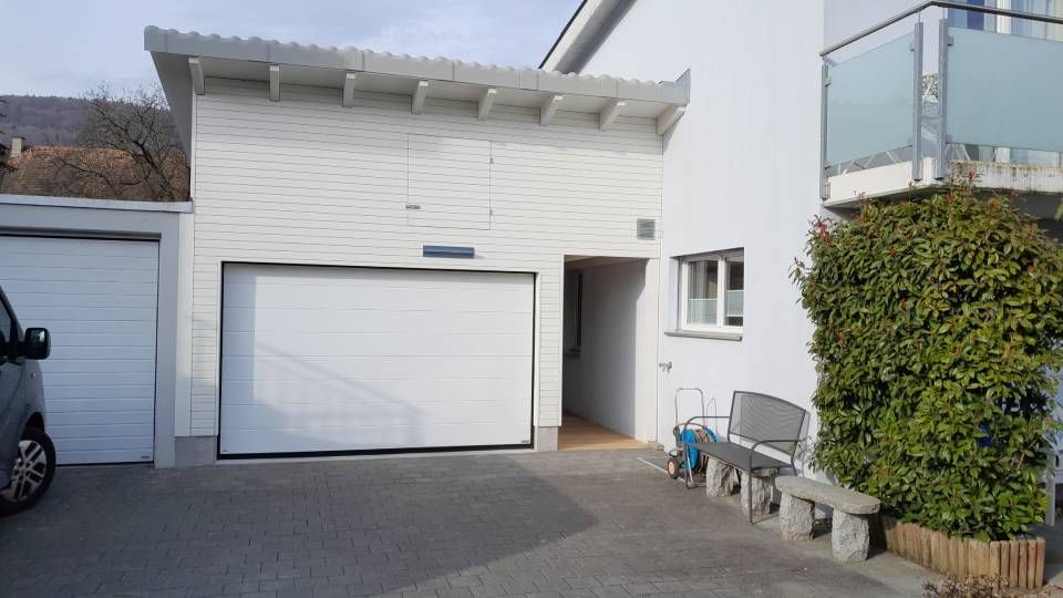 Aussenansicht eines weissen Hauses mit Garagentor, kleinem Eingang und von einer Hecke gesäumtem Seitenweg unter strahlend blauem Himmel