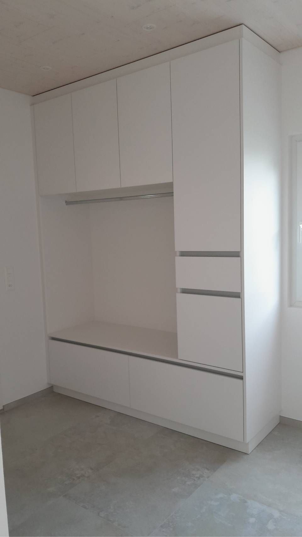 Ein weisser Einbauschrank und Aufbewahrungsschränke in einem kahlen Zimmer, rechts davon ein Fenster.