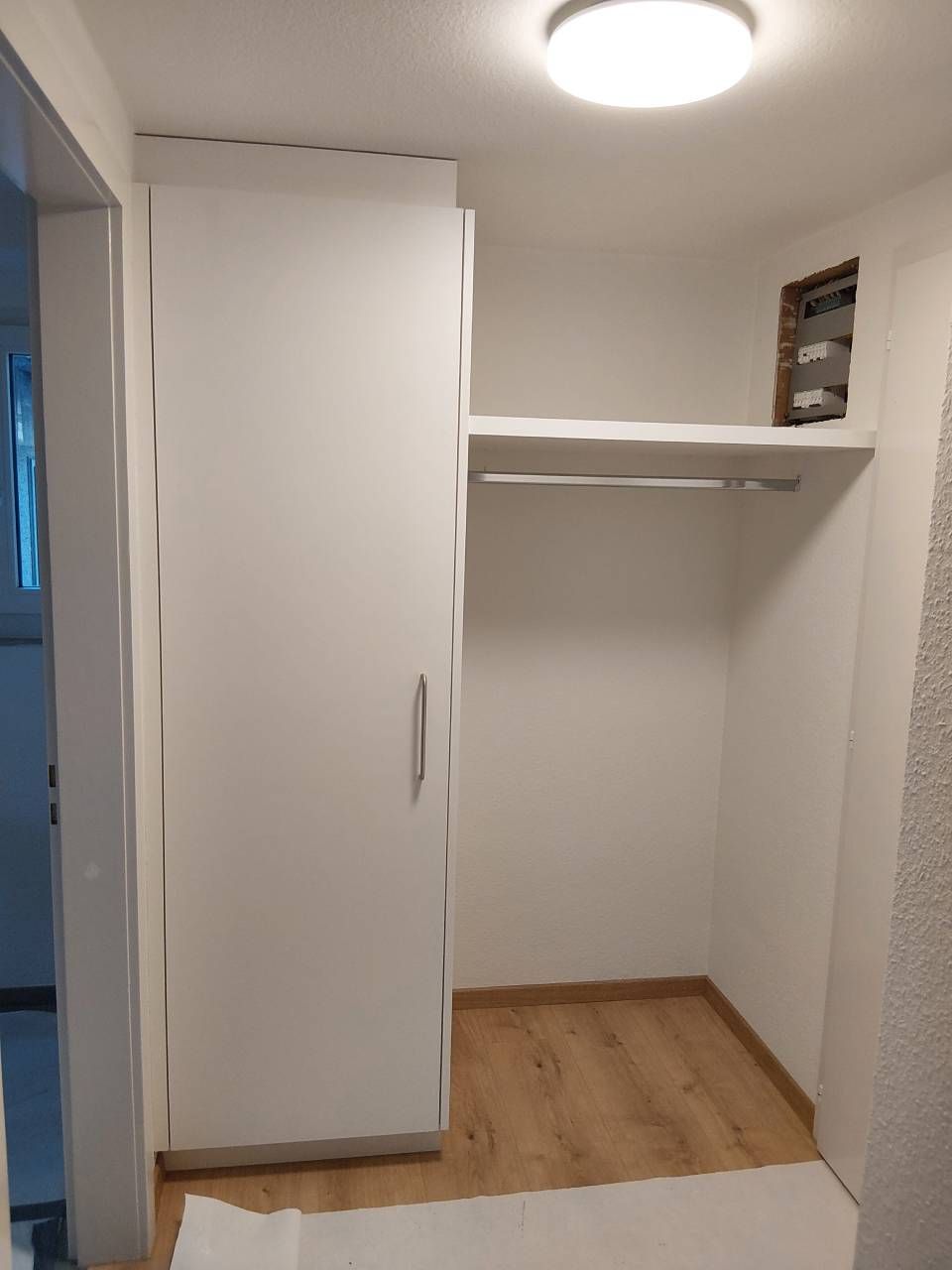 Kleiner weisser Schrank mit Holzboden und einem Regal unter einer Deckenleuchte