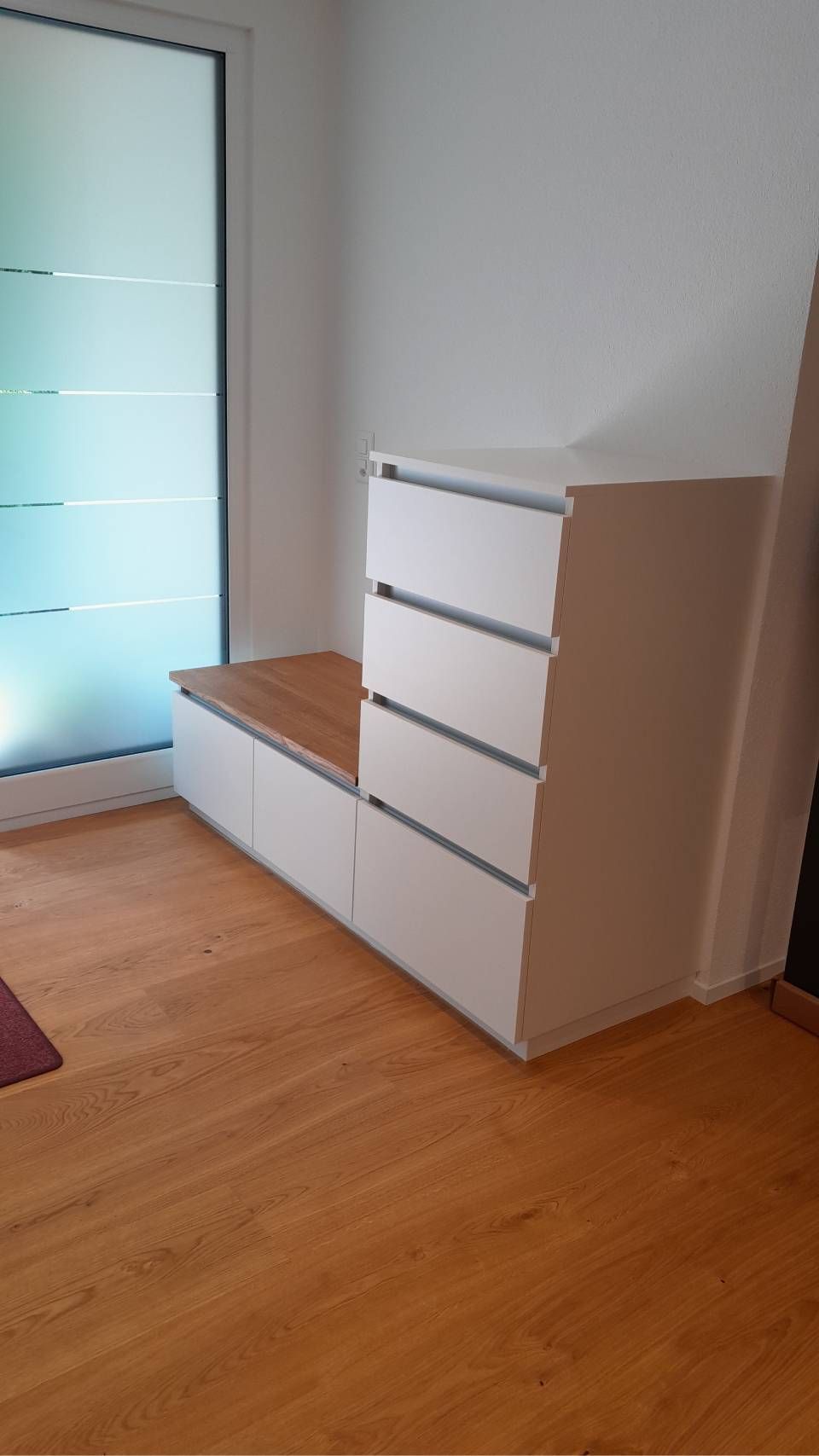 Weisse Kommode mit Holzplatte und Bank in einem hellen Zimmer mit Holzboden und Milchglastür