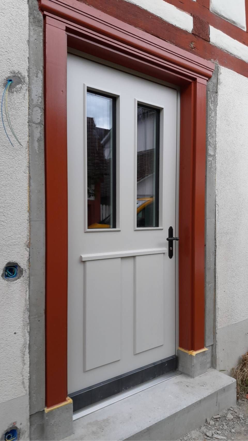 Weisse Eingangstür mit Glasscheiben und schwarzem Türgriff an einer rot umrandeten Gebäudefassade.