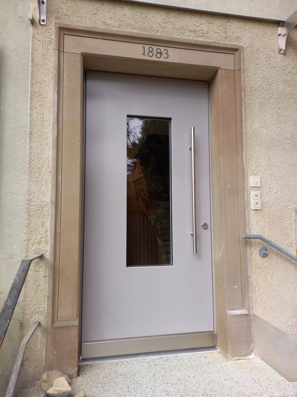 Moderne graue Eingangstür mit schmalem Fenster und Griff, eingelassen in eine beigefarbene Gebäudefassade mit der Hausnummer 1883