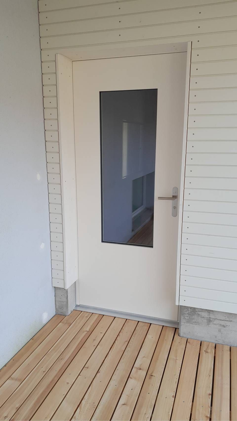 Weisse Aussentür mit schmalem Glasfenster an einer kleinen Holzveranda mit weisser Verkleidung.