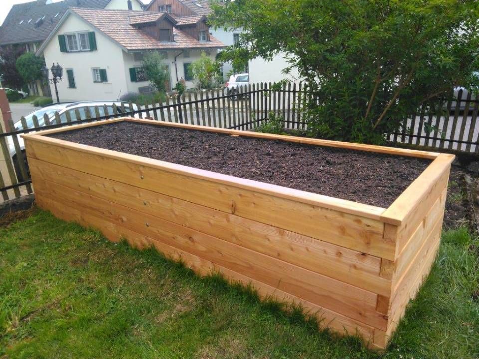 Ein hölzernes Hochbeet, gefüllt mit Erde, steht in einem grasbewachsenen Hinterhof in der Nähe eines Zauns und von Häusern.