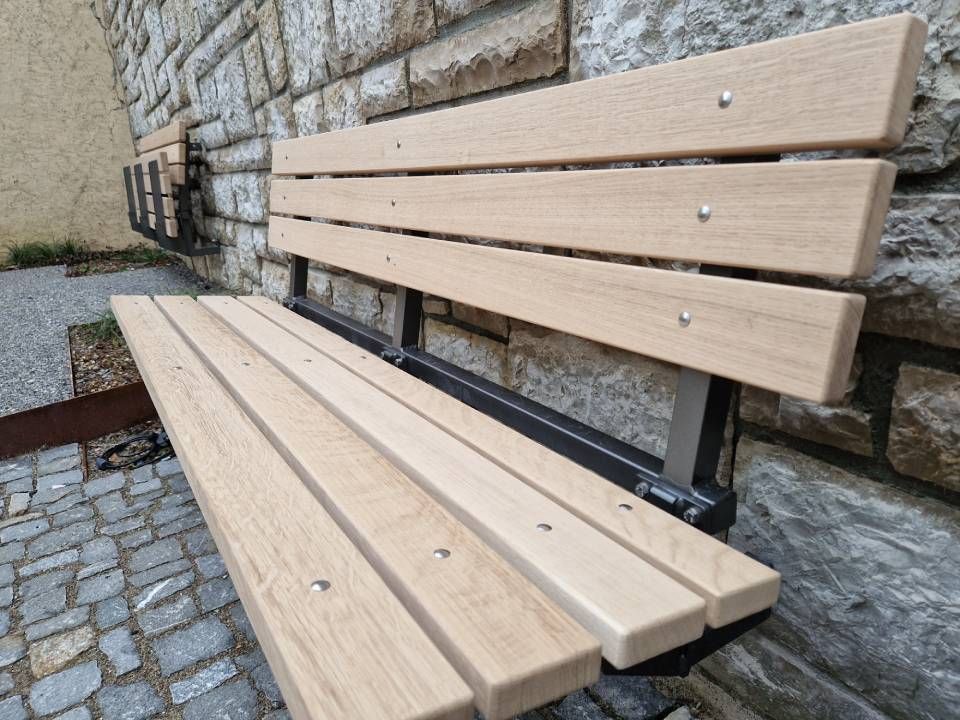 Holzbank mit schwarzen Metallstützen neben einer Steinmauer auf einem Kopfsteinpflasterweg
