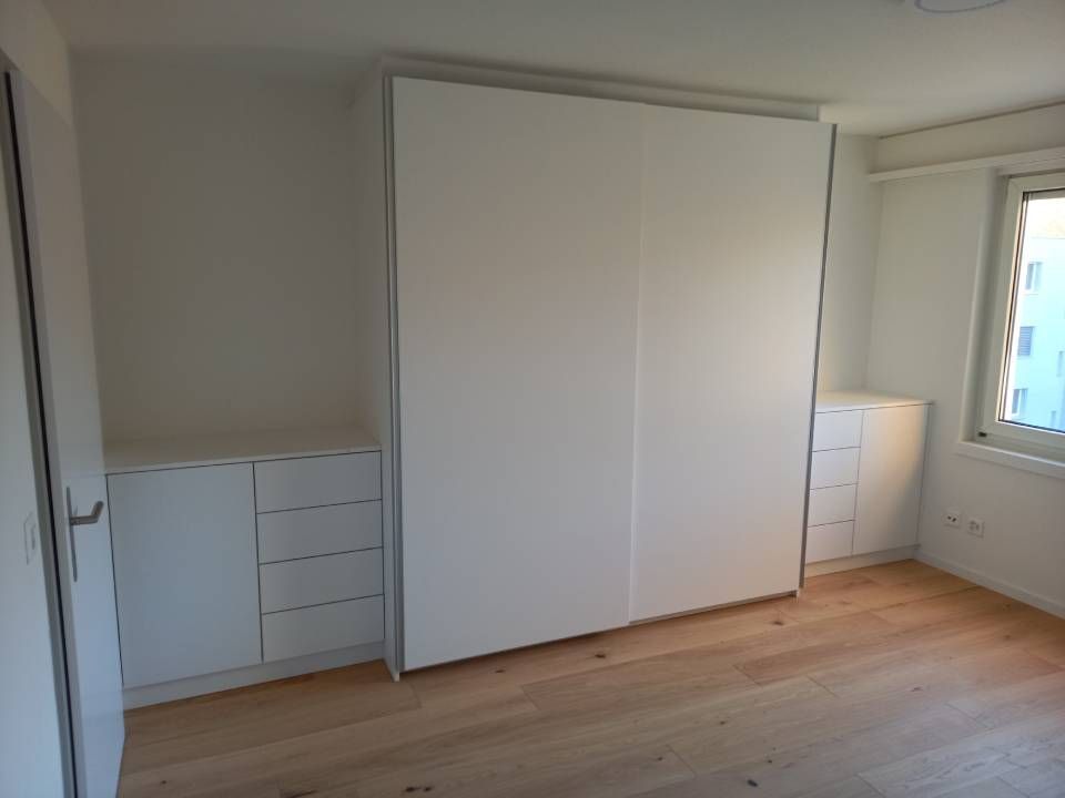 Leeres weisses Schlafzimmer mit Holzboden, Einbauschrank, Kommoden und einem Fenster auf der rechten Seite.