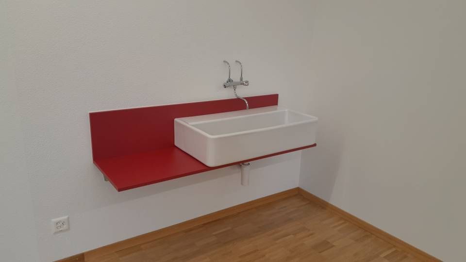 Weisses Wandwaschbecken mit rotem Spritzschutz in einem spärlich eingerichteten Raum mit Holzboden