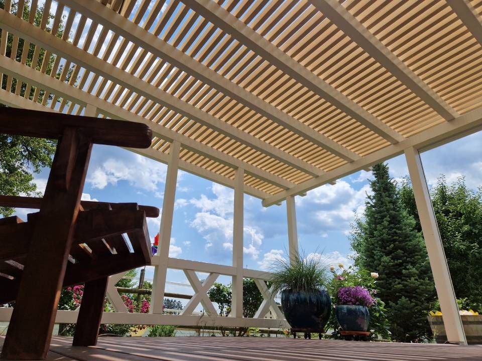 Überdachte Terrasse mit Lattenpergola, weisser Bank und Topfblumen mit Blick auf einen sonnigen Garten