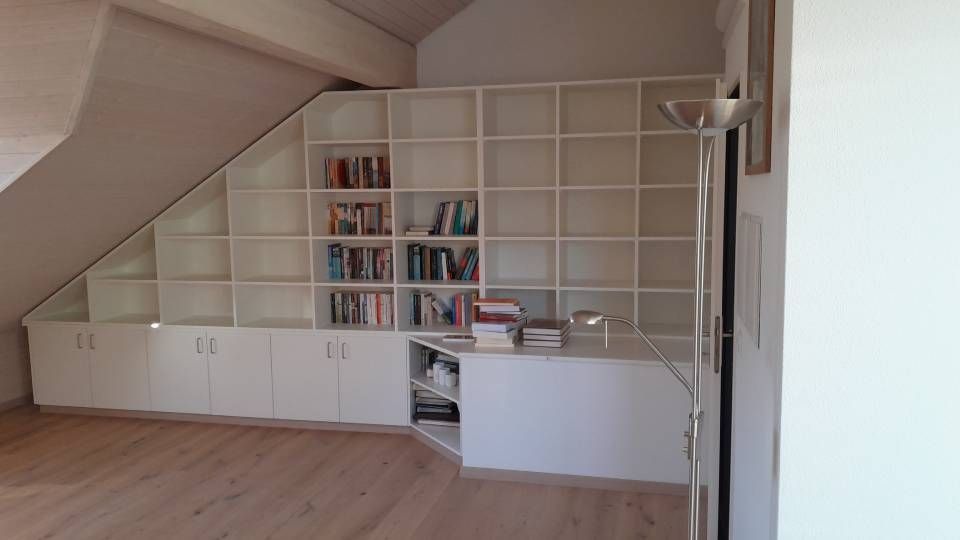 Kleines Dachzimmer mit eingebauten weissen Schränken, Regalen, Büchern und einem Schreibtisch unter einer Dachschräge.