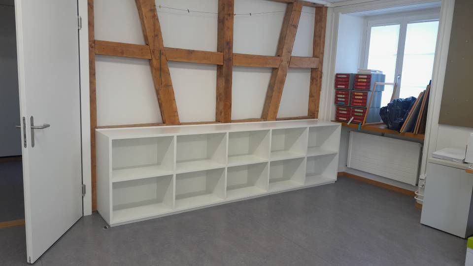 Ein leerer, weisser Raum mit eingebauten Regalfächern, grossen Fenstern und Holzbalken an der Wand.