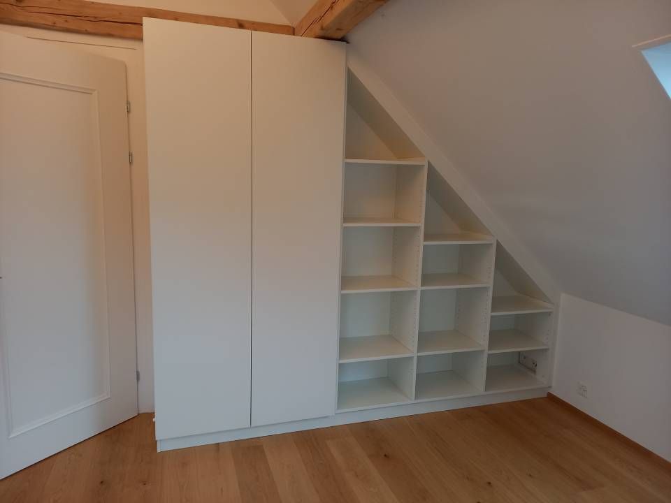 Weisse Einbauregale und ein hoher Schrank in einem kleinen Dachzimmer mit Dachschräge und Holzboden