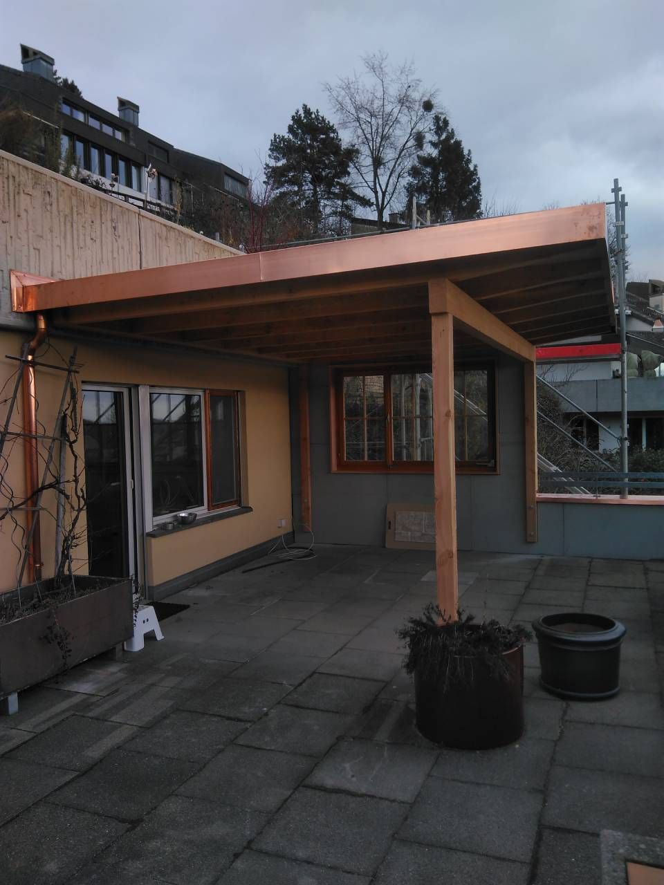 Hinterhofterrasse mit hölzernem Pergola-Dach, Topfpflanzen und grauen Pflastersteinen an einer Hauswand