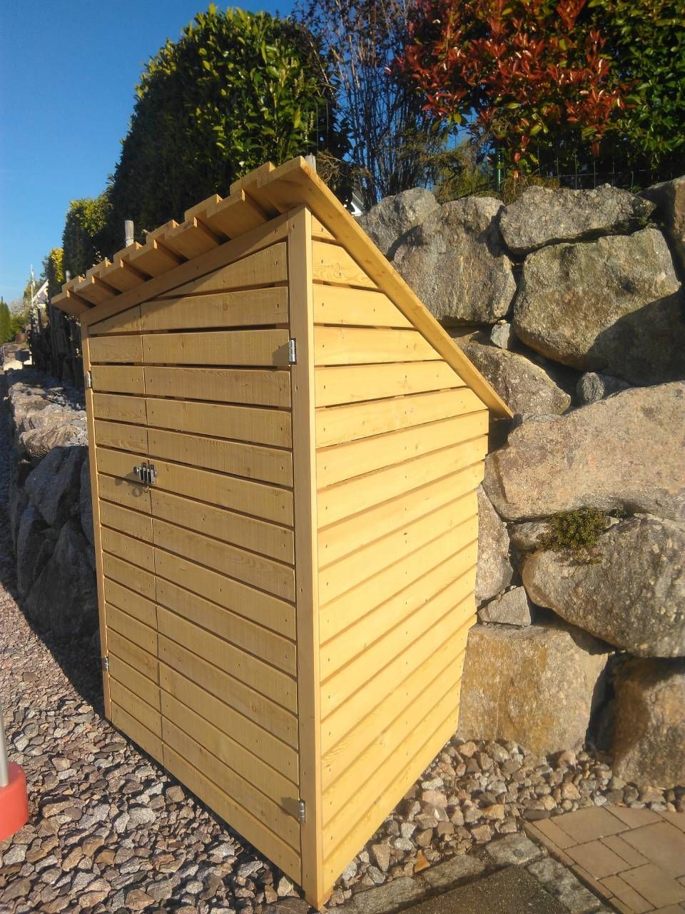 Ein hölzerner Geräteschuppen im Freien neben grossen Steinen auf einer Kiesterrasse, dahinter blauer Himmel und Bäume