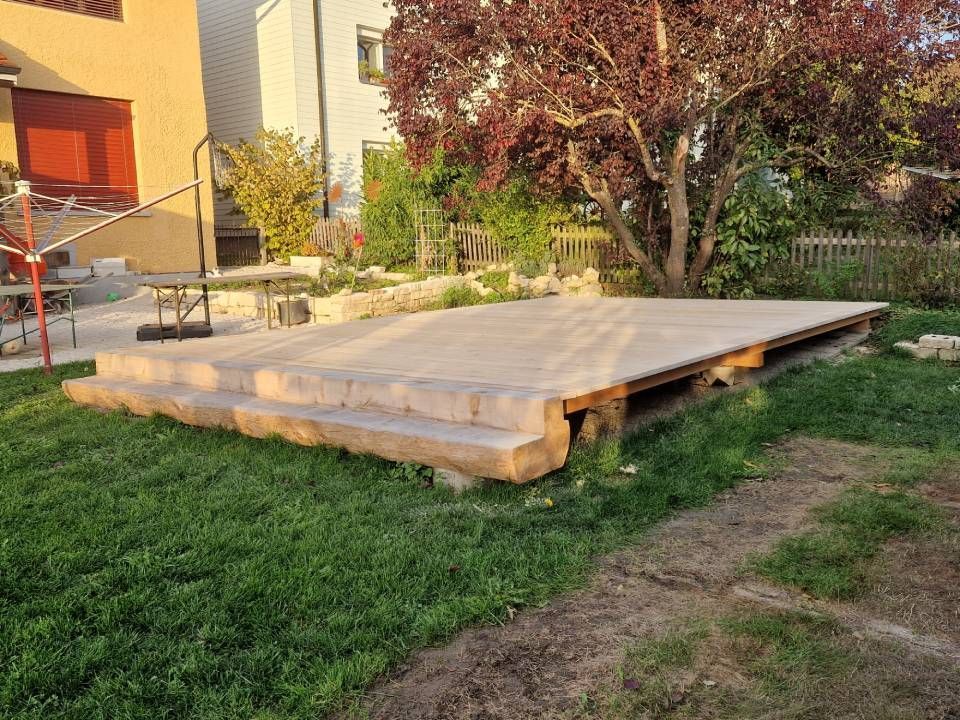 Eine erhöhte Holzplattform in einem grasbewachsenen Hof neben einem Haus, mit Stufen und nahegelegenen Bäumen.