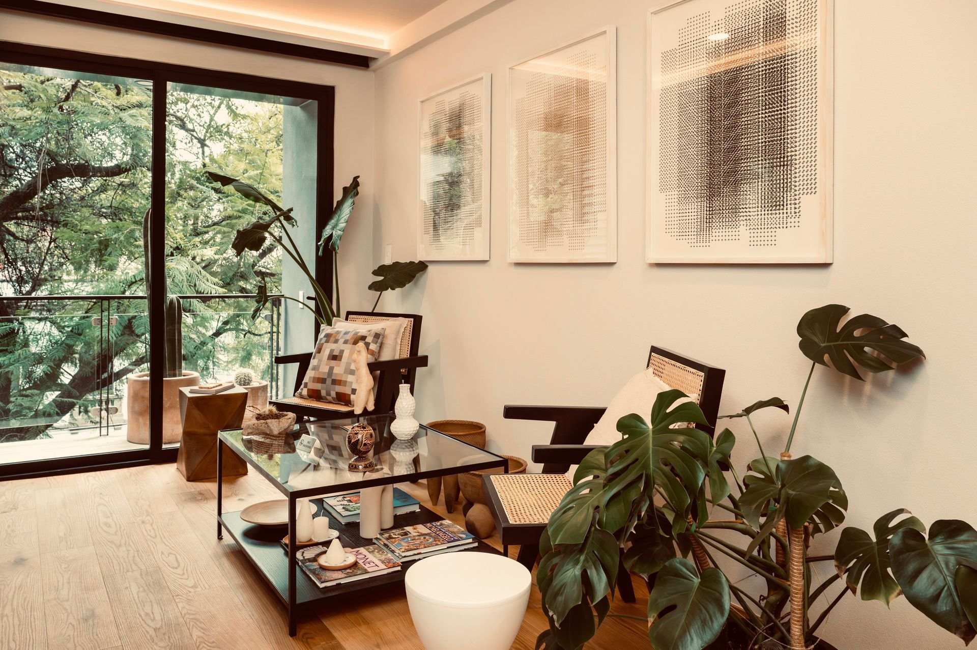 Living room with glass-top coffee table, two chairs, and large Monstera plant. Balcony with trees visible. Art on wall.