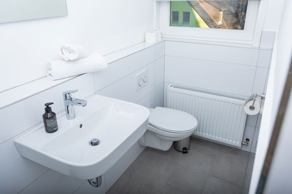 Ein modernes weißes Badezimmer mit wandmontiertem Waschbecken, Toilette, Heizkörper und grauem Fliesenboden.