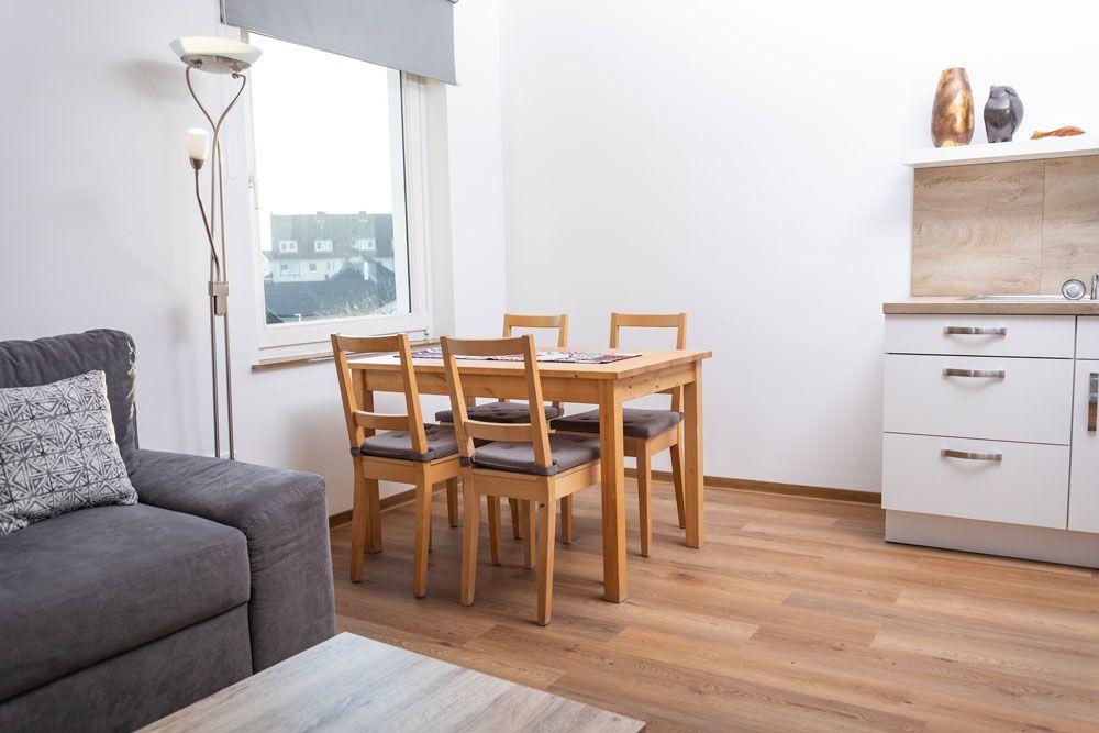 Ein helles Zimmer mit einem Esstisch aus Holz und vier Stühlen, einem grauen Sofa, einer Stehlampe und einem weißen Küchenschrank.