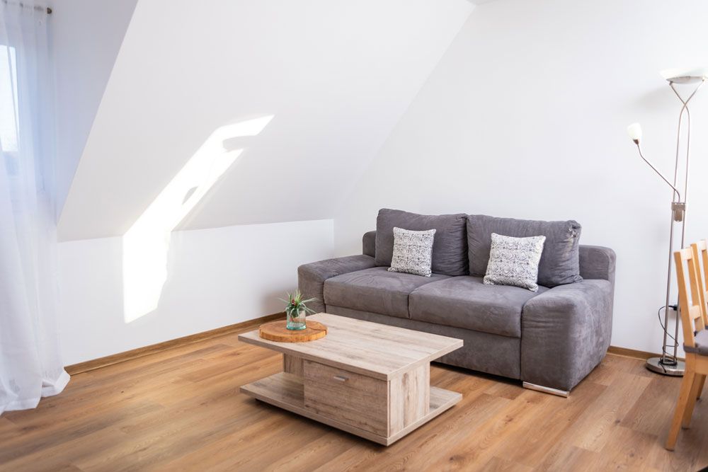In einem hellen, dachgeschossartigen Zimmer mit Holzboden und einem hellen Couchtisch aus Holz steht ein graues Sofa mit gemusterten Kissen.