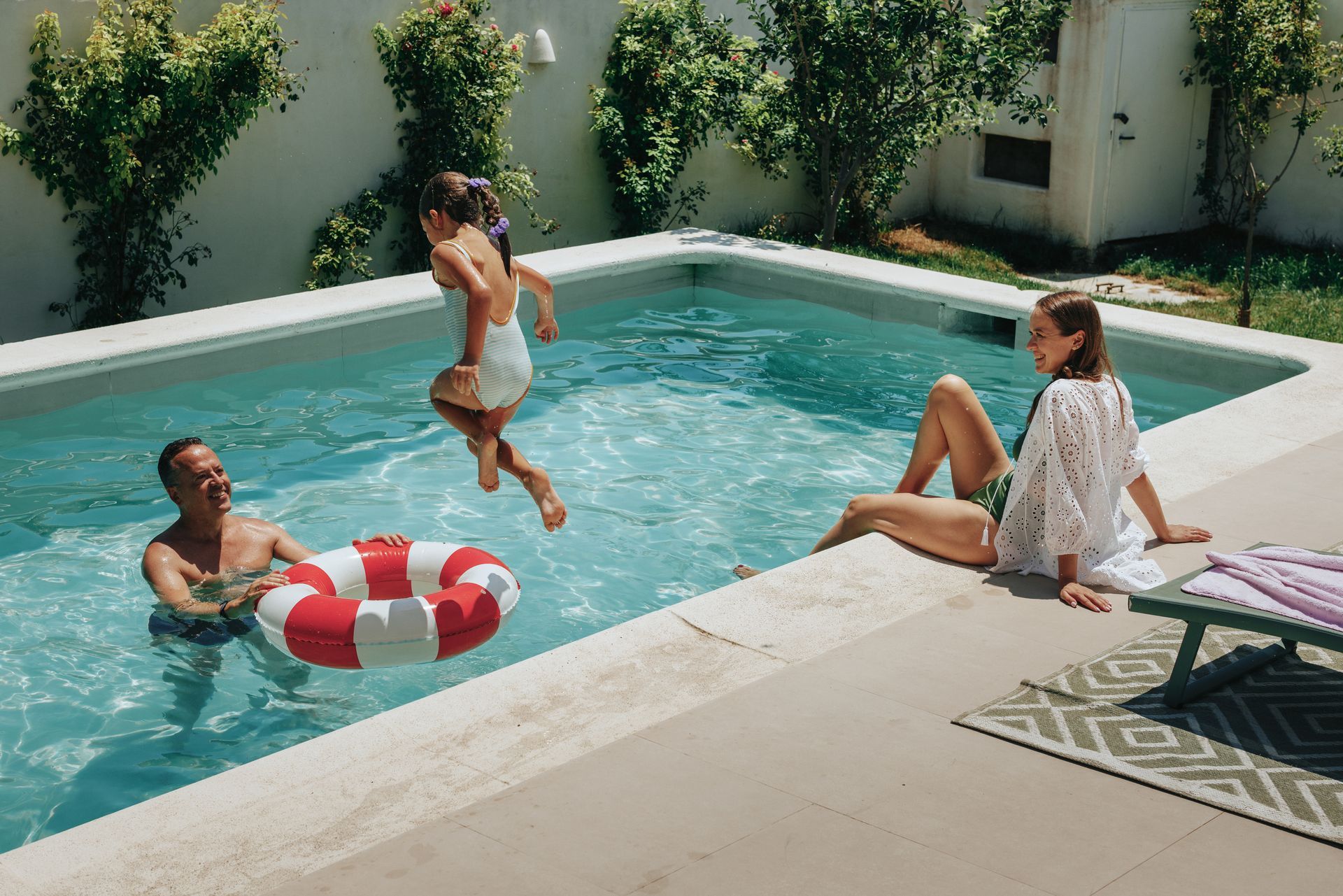 Trois personnes se détendent au bord d'une piscine dans un jardin, l'une d'elles plongeant à proximité d'une bouée rouge et blanche.