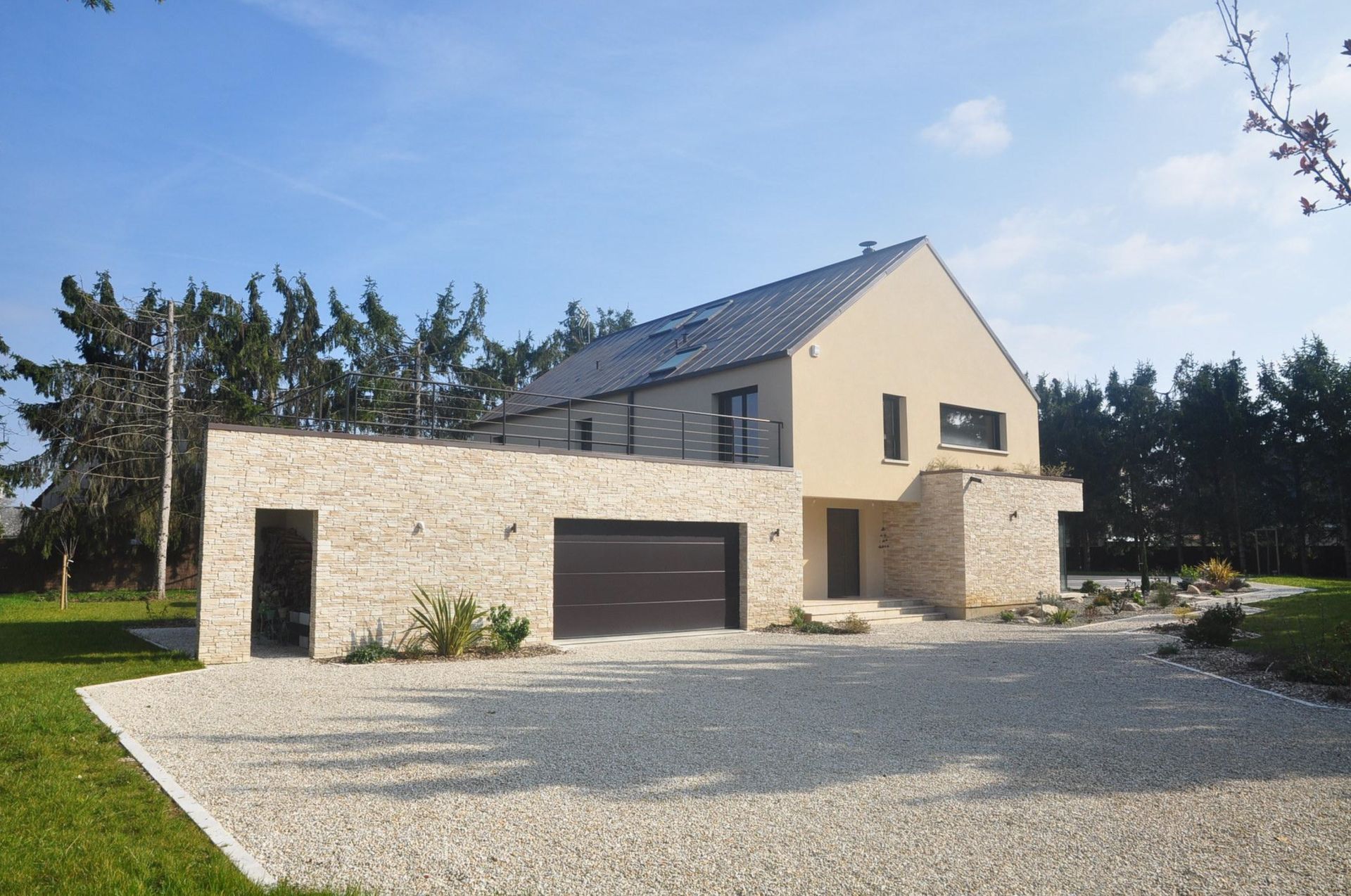 Maison beige moderne avec garage en pierre, allée en gravier et arbres sous un ciel bleu