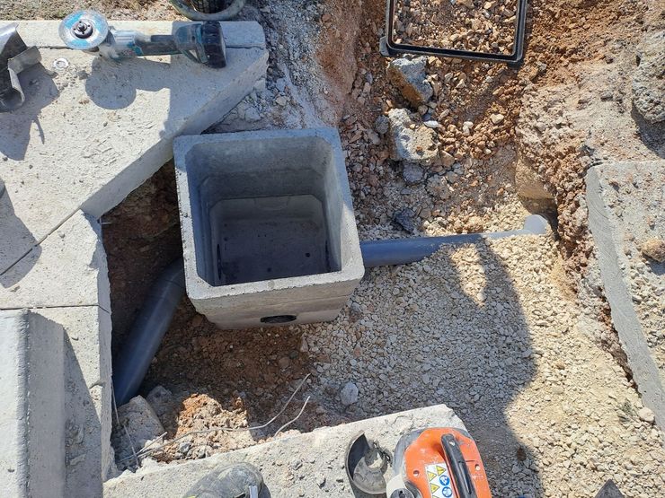 Fosse utilitaire ouverte avec chambre carrée en béton, tuyaux et outils dans une zone de construction en terre