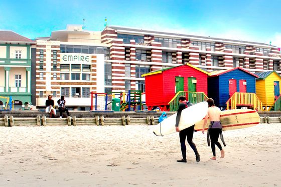 Two people carrying surfboards on a beach in front of the Empire Building in Muizenberg, Cape Town