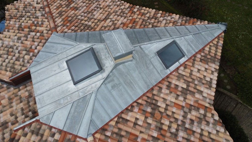 Vue aérienne d'un toit en panneaux de zinc avec une cheminée centrale et deux puits de lumière, sur fond de toit en tuiles de terre cuite.